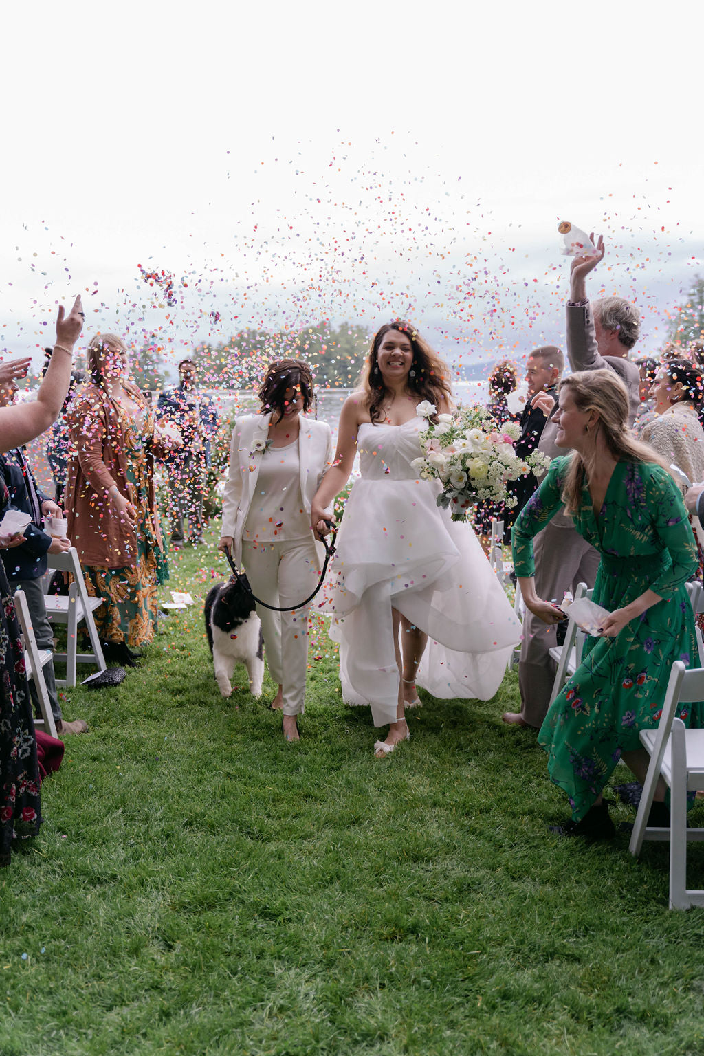 Two bride walking down the aisle at Migis Lodge in Casco, Maine with confetti being thrown, surrounded by guests on a grassy outdoor setting