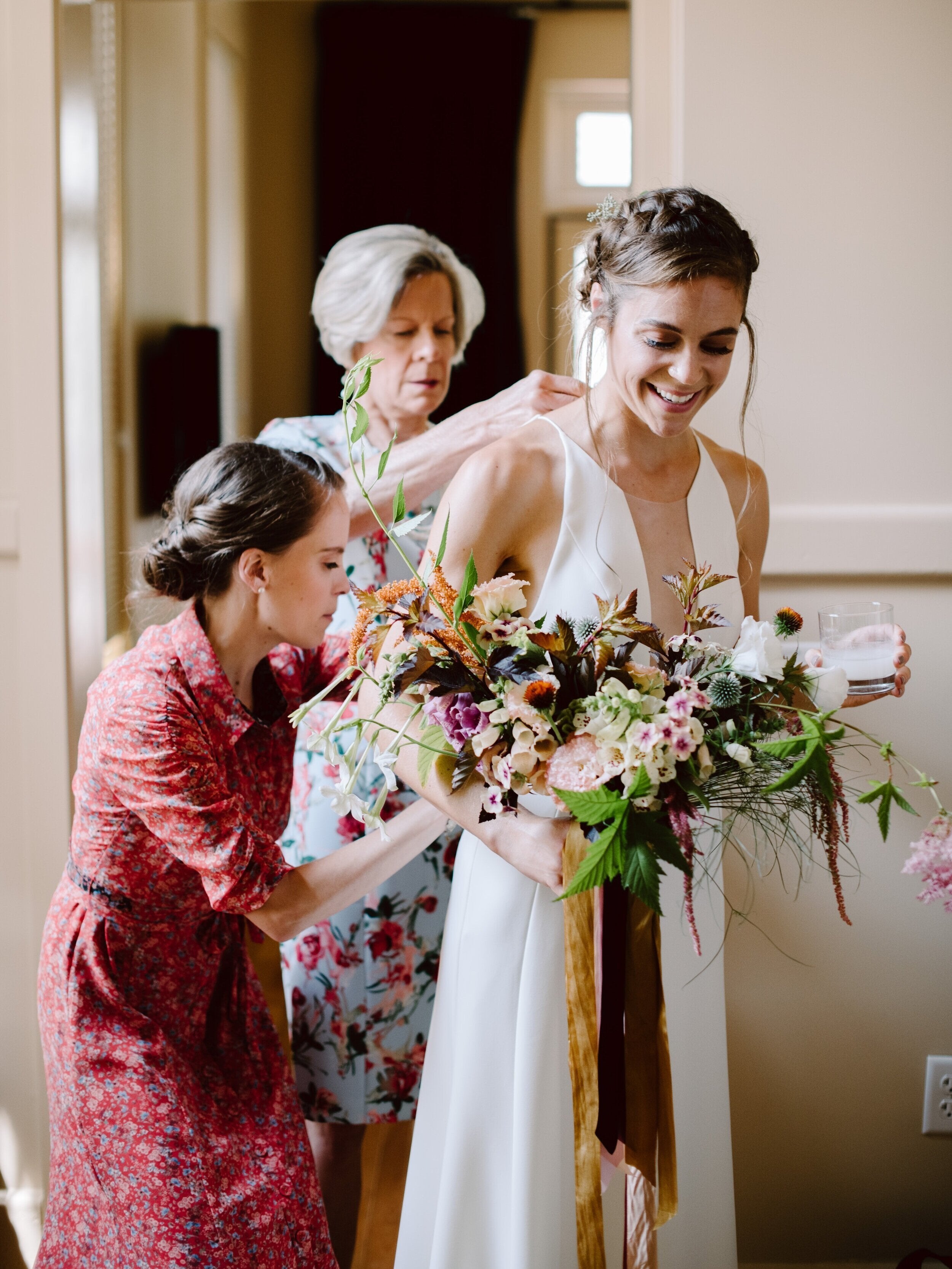 Woman in a white dress holding a bouquet, with two other women assisting her.