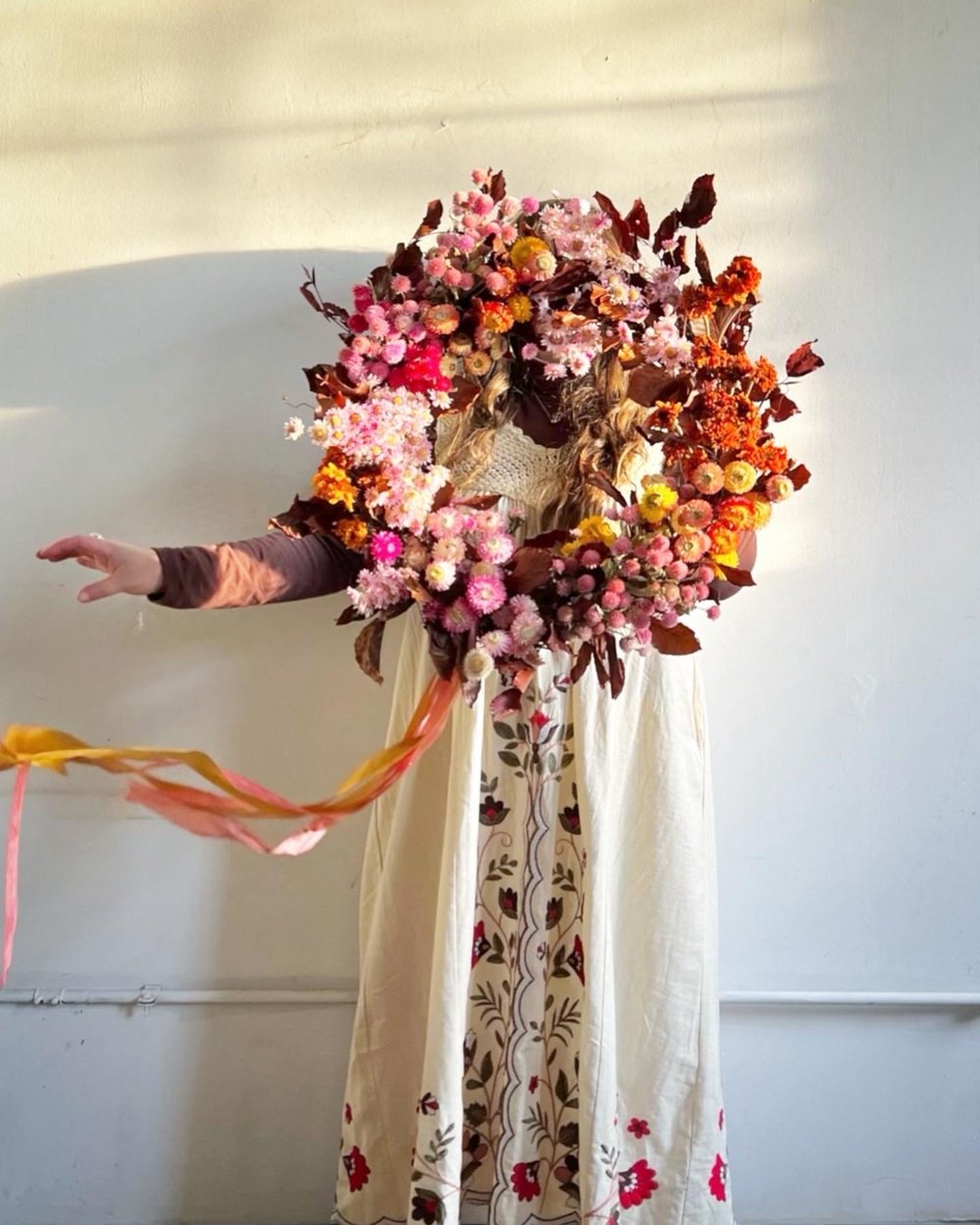Person in flowy white dress holding floral wreath with colorful flowers against a white background
