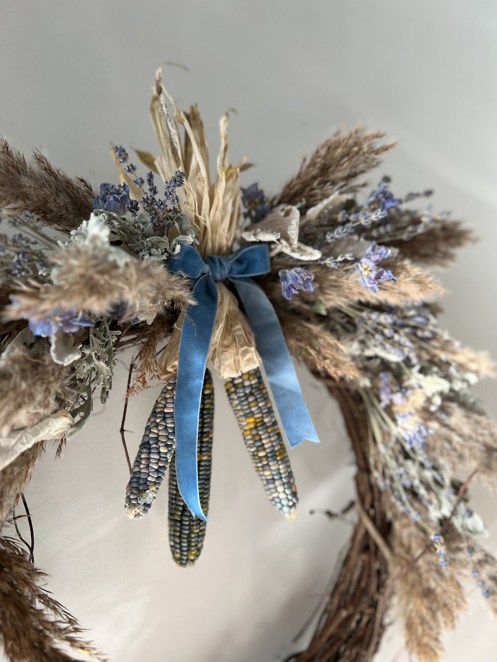 Decorative wreath with dried flowers, corn cobs, and a blue ribbon on a neutral background
