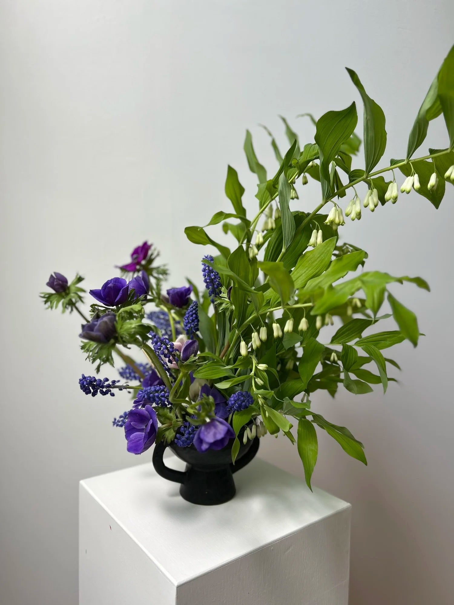 Bouquet of purple flowers and green leaves in a black vase on a white pedestal against a light gray background
