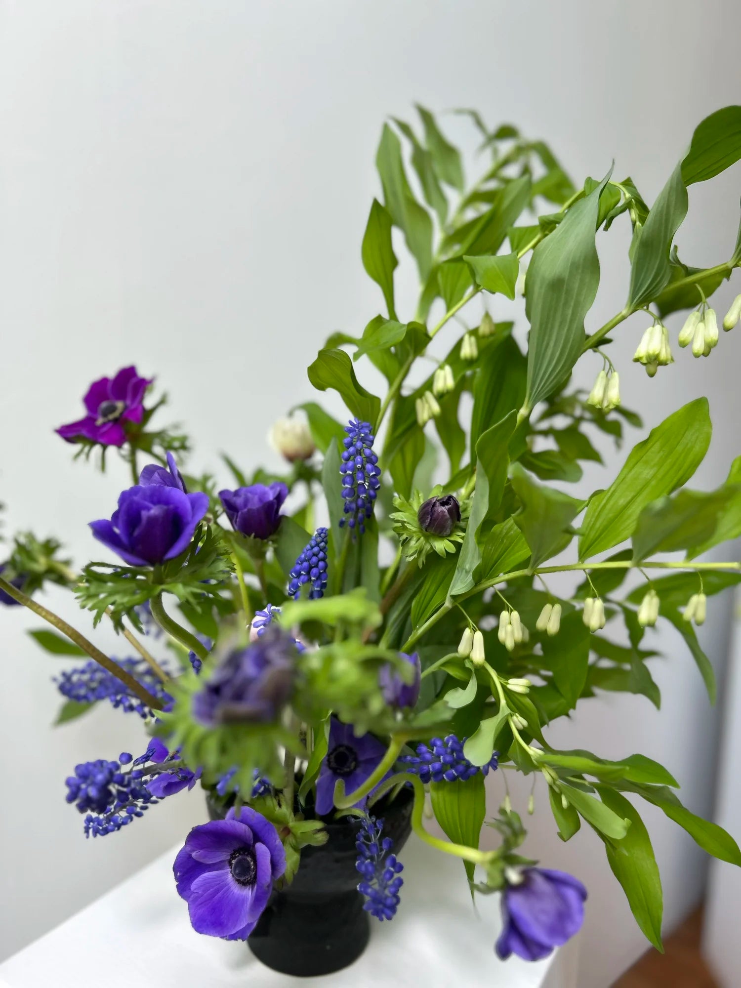Bouquet of purple flowers and green leaves in a black vase on a white surface.