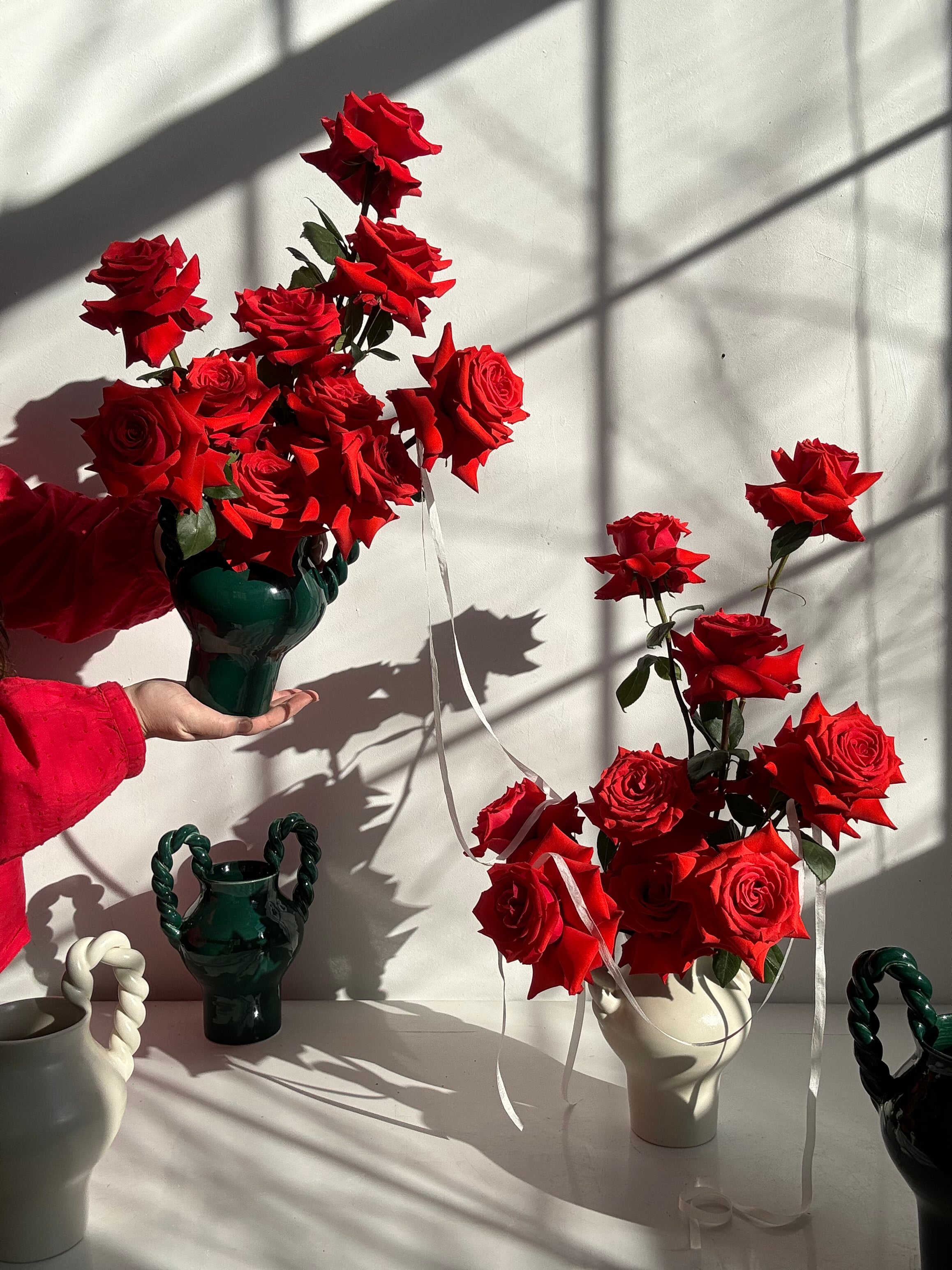 Red roses tied with long silk ribbons arranged in handmade Maine made ceramic vases on a white surface with shadows cast by hands and objects.