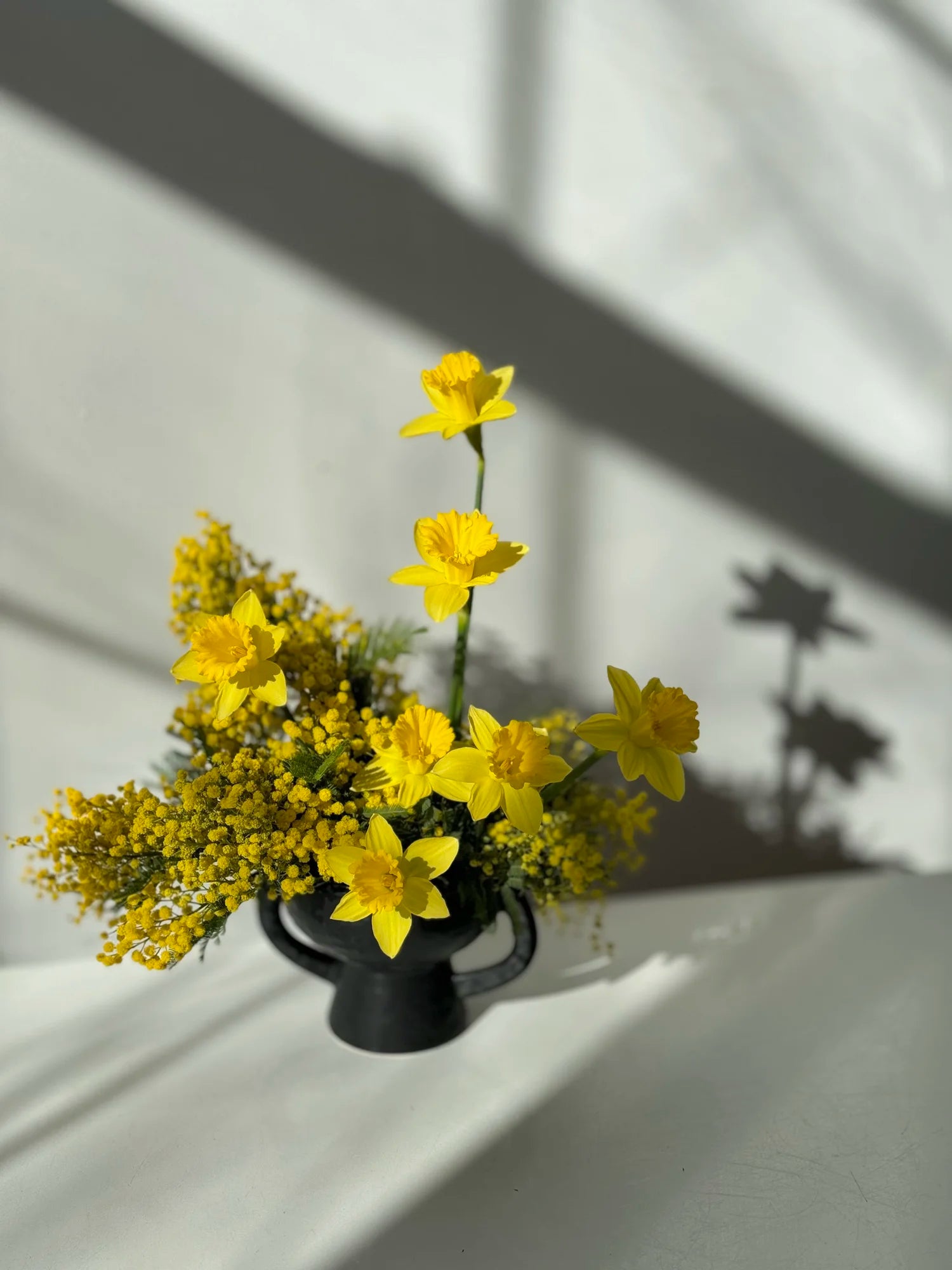 Yellow flowers in a black vase on a white surface with shadows.