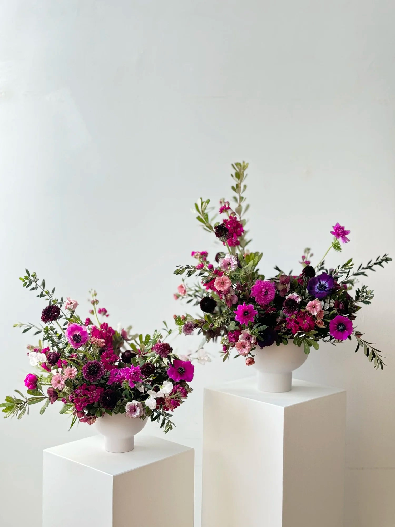 Two floral arrangements in white vases on white pedestals against a light gray background