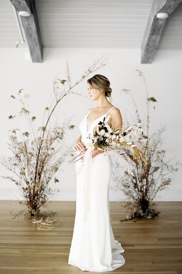 Woman in a white dress holding a bouquet of flowers in a minimalistic setting with dried plants.