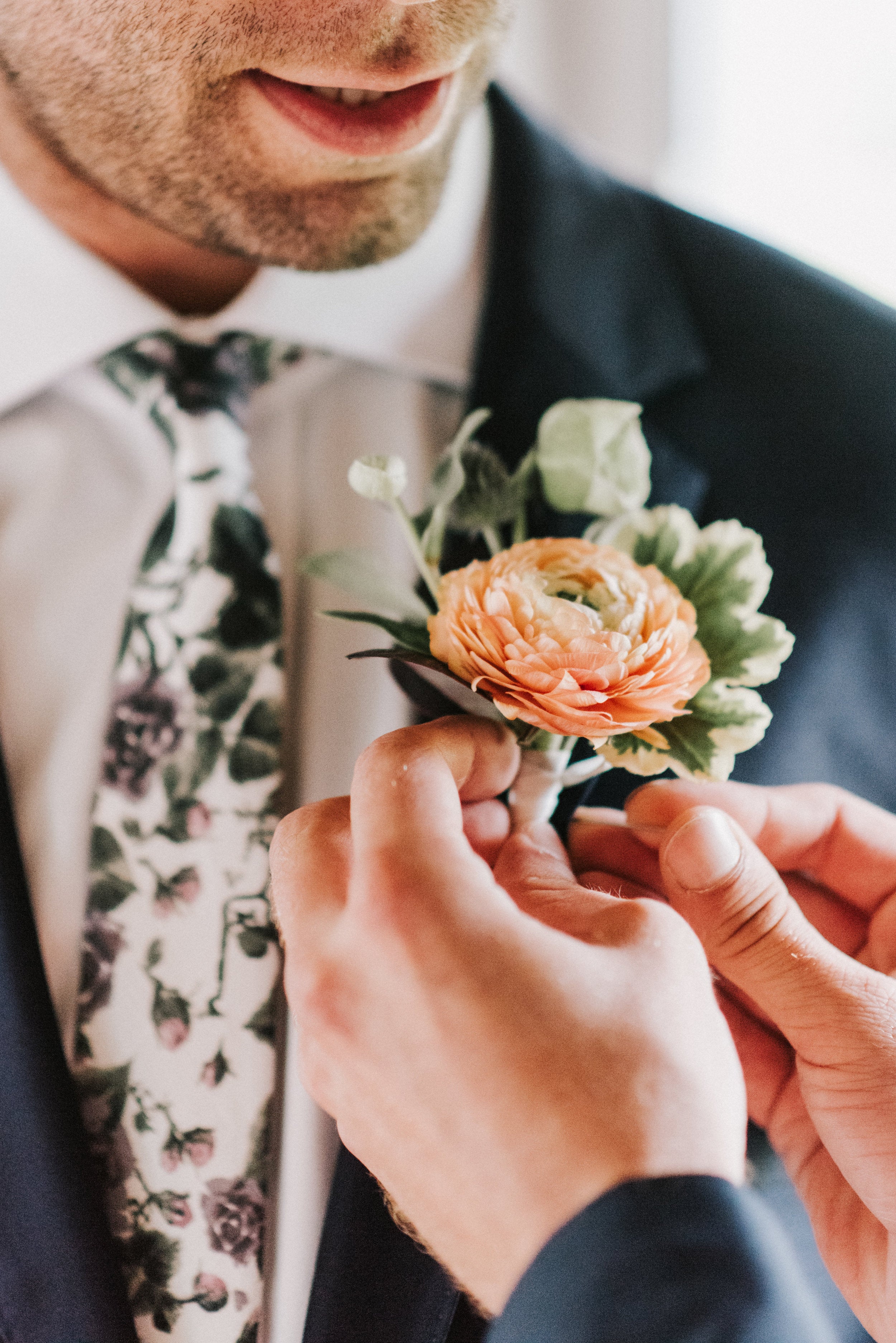 Person adjusting a floral boutonniere on another person's suit jacket.