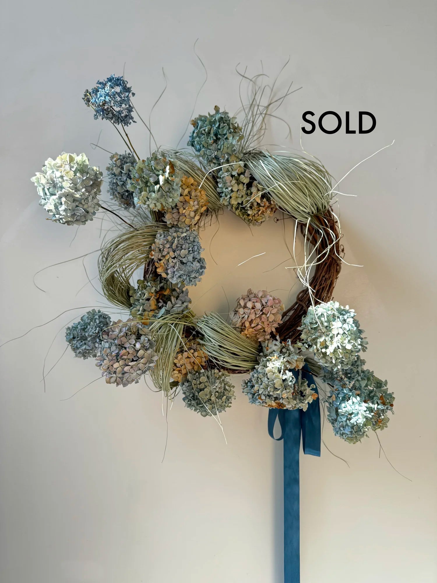 Decorative wreath with flowers and a blue ribbon on a plain background, with 'SOLD' text above.