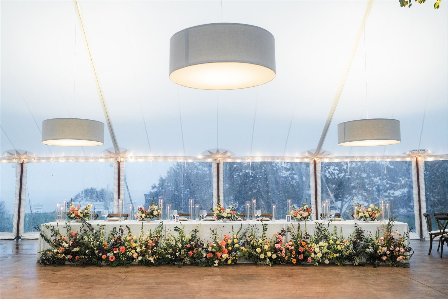 Long table set for a formal event with floral decorations and large hanging lights in a room with large windows.