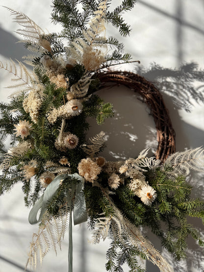 Decorative wreath with greenery, flowers, and a brown ribbon on a neutral background