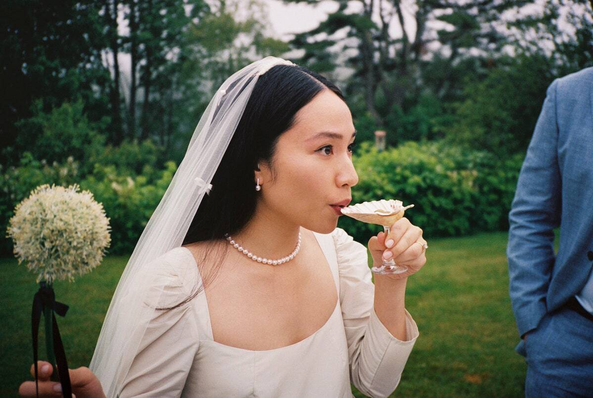 Bride sipping from shell-shaped glass holding single allium with black bow