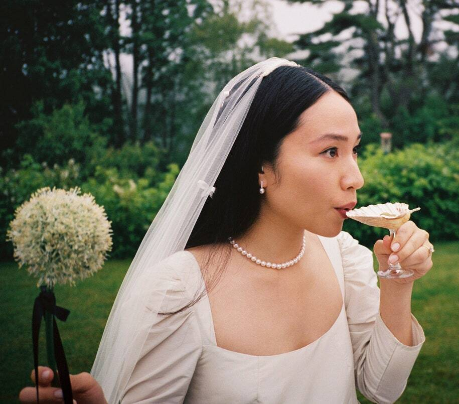 Woman in a white bridal outfit holding a bouquet and a glass of champagne outdoors.