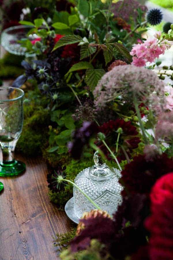 Decorative table setting with flowers, a glass bottle, and green glasses on a wooden surface.