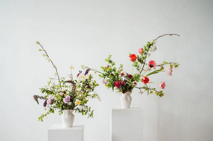 Two flower arrangements in white vases on white pedestals against a light gray background