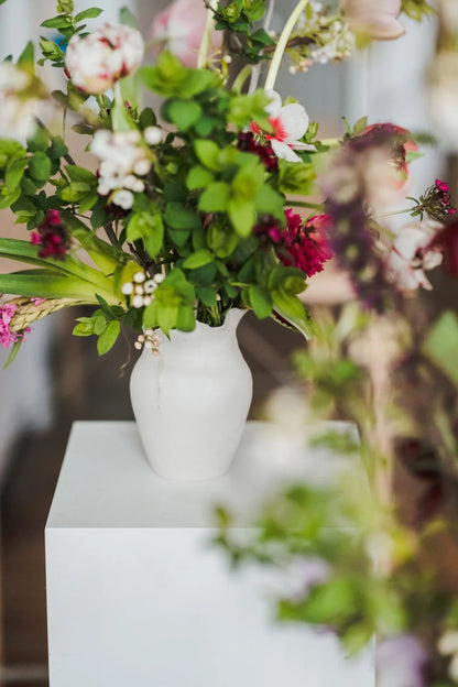 White vase with flowers on a white pedestal against a blurred floral background