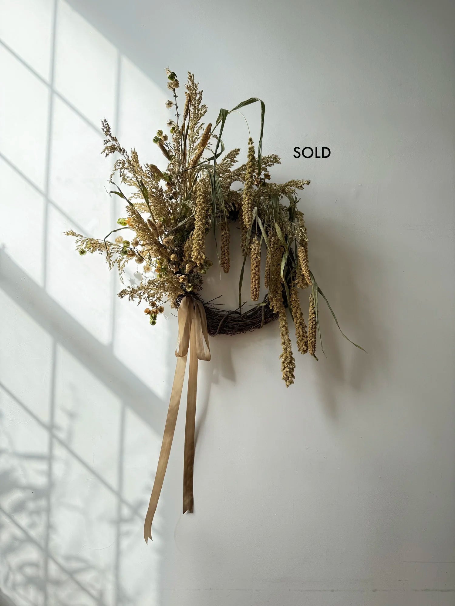 Decorative wall piece with dried plants and a sold label on a white wall.