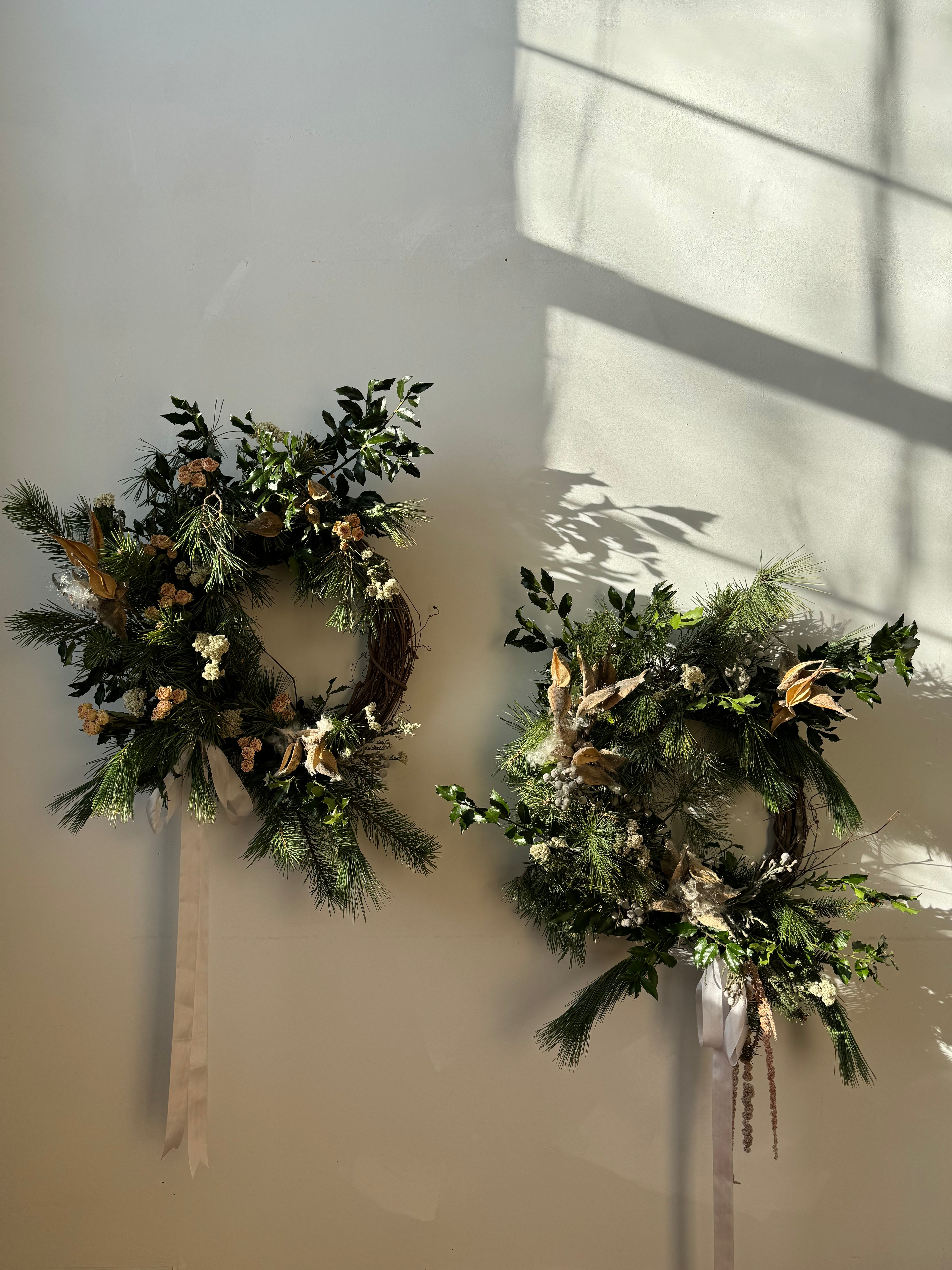 Two holiday evergreen wreaths with floral materials and long bows on a white wall.
