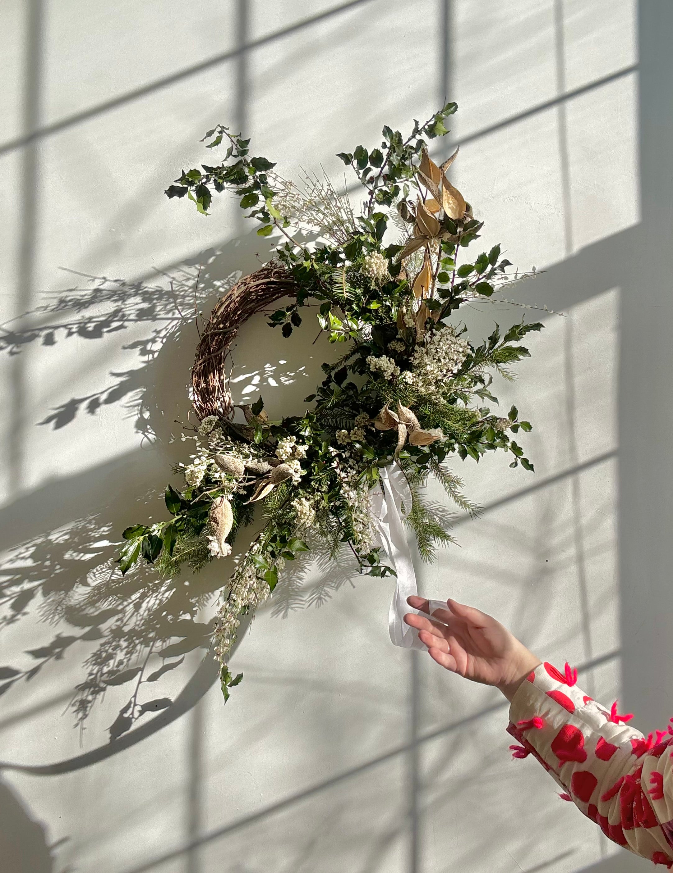 Maine florist's hand holding a decorative  evergreen and floral wreath against a white wall with shadows.
