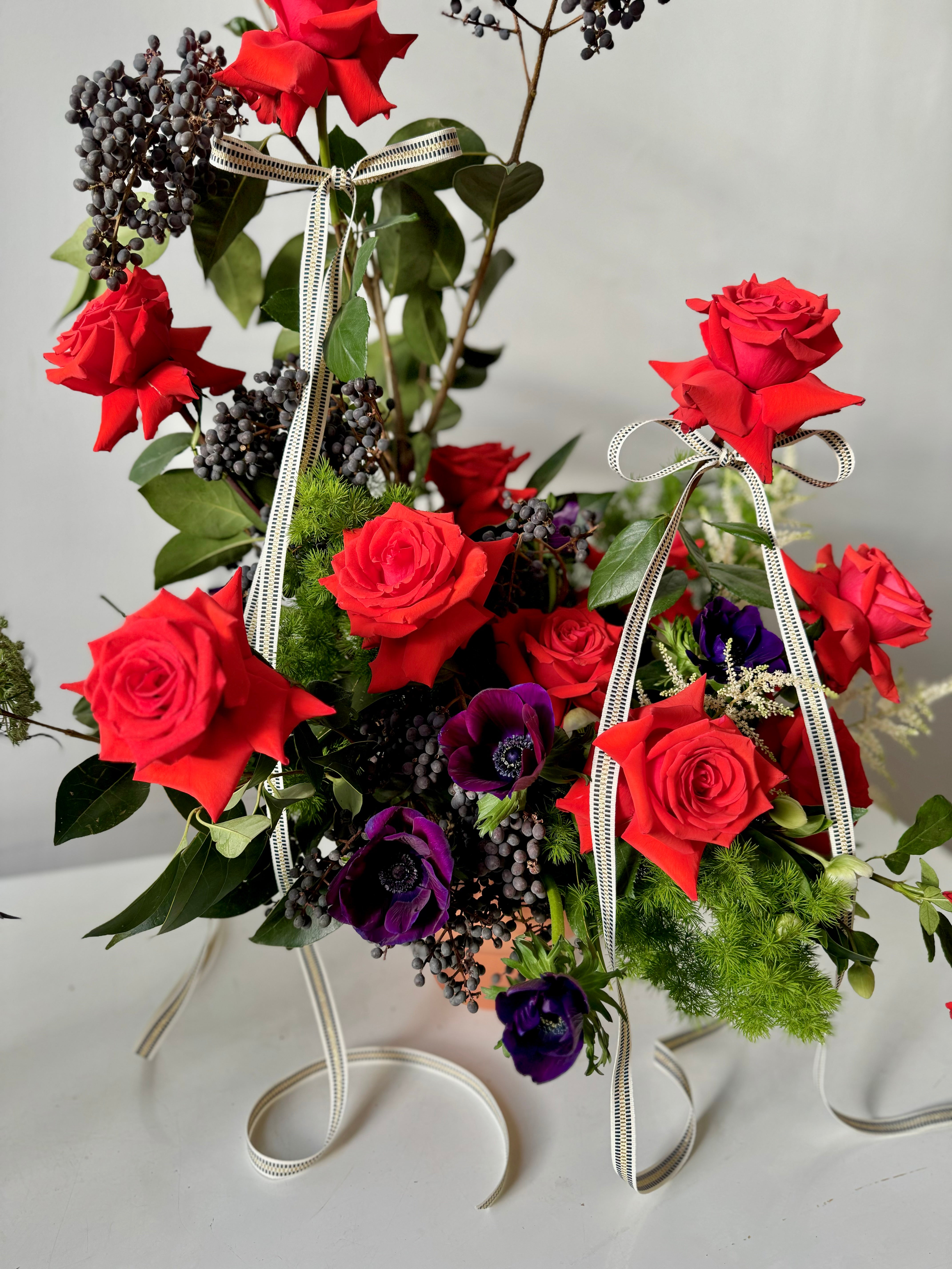 Holiday floral arrangement with red roses , privet berries, and purple anenomes tied with patterned ribbons on a white table against a gray background