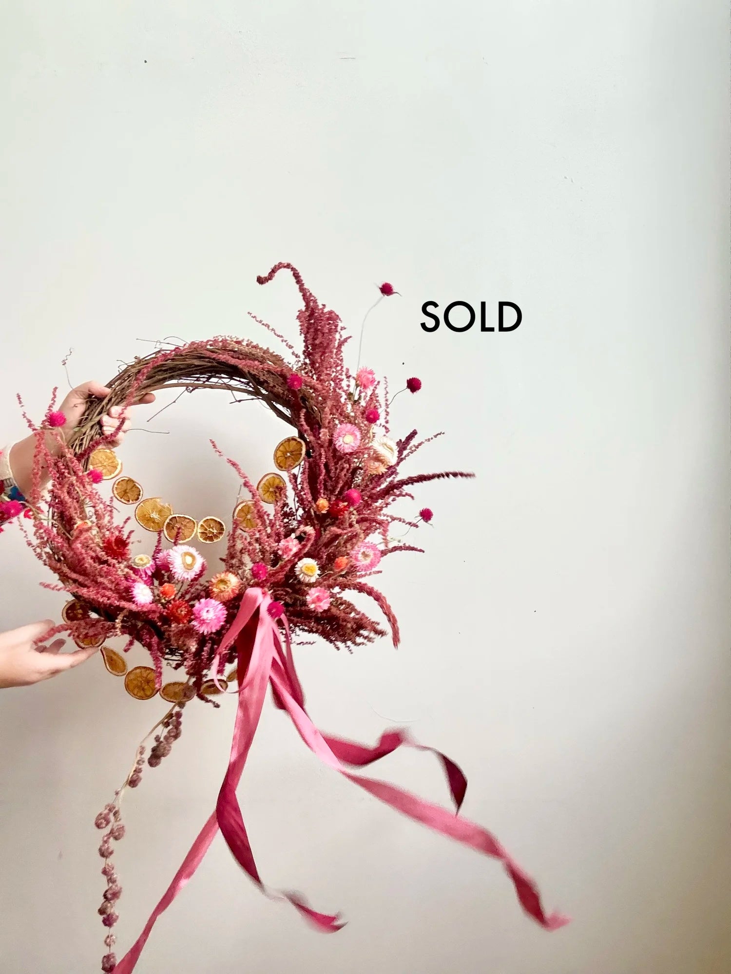 Decorative wreath with pink ribbons and beads on a plain background, with 'SOLD' text.