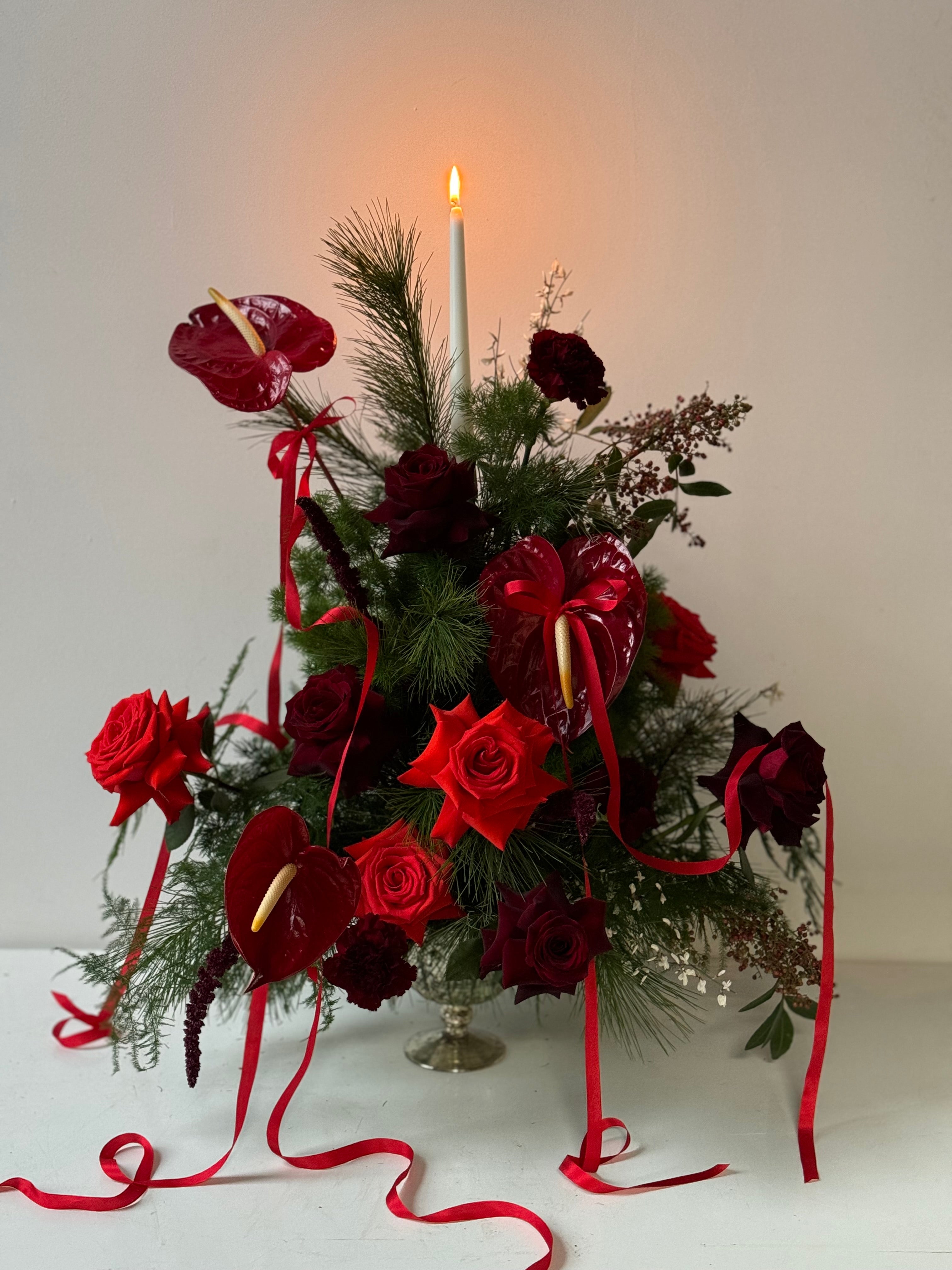 Decorative floral arrangement and topiary with red roses, anthurium, evergreens in a silver compote with a tall candle on a white background