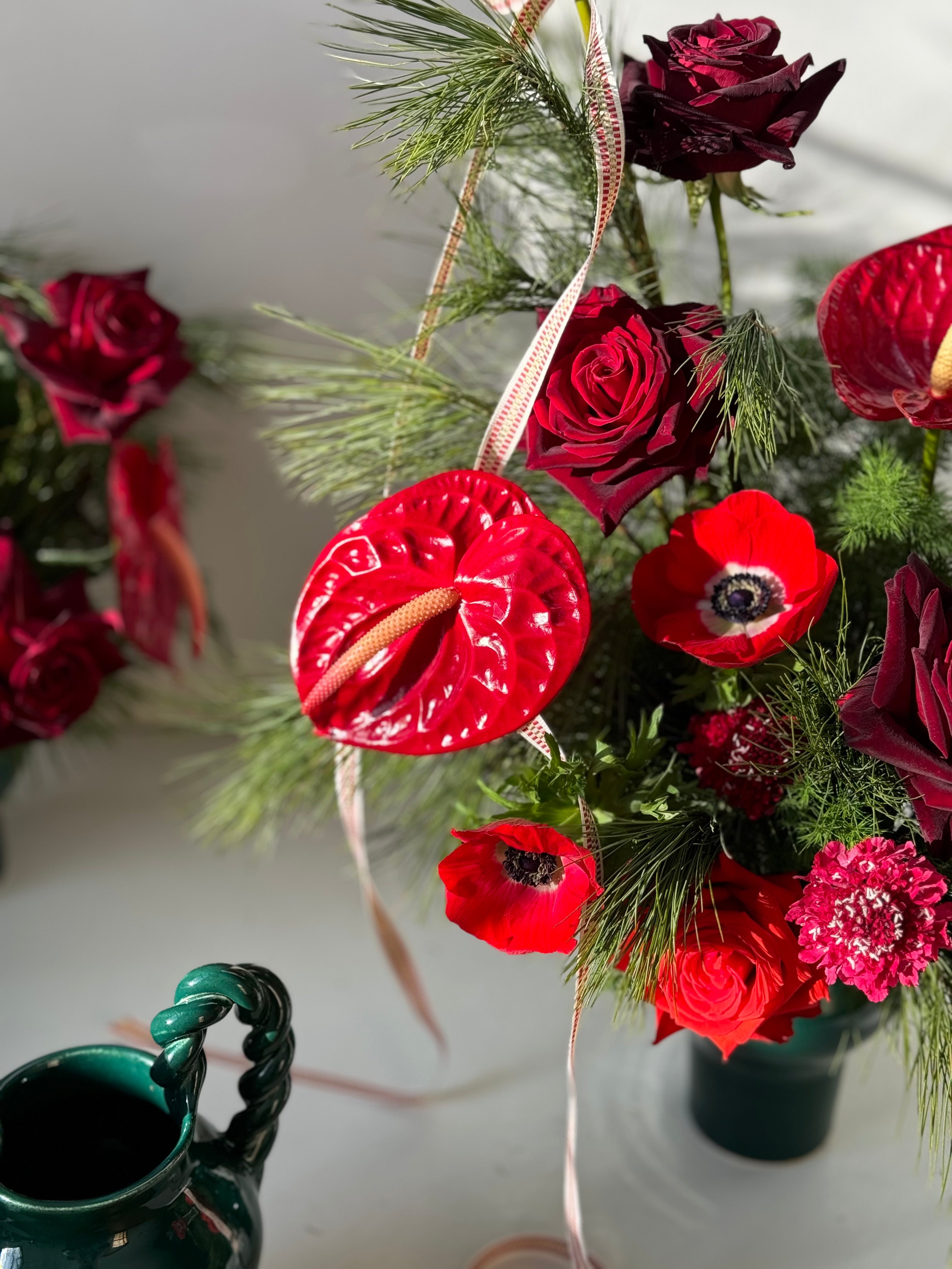 Decorative holiday arrangement of red anthurium, anenomes, and evergreen in a green handmade in Maine vase on a light background