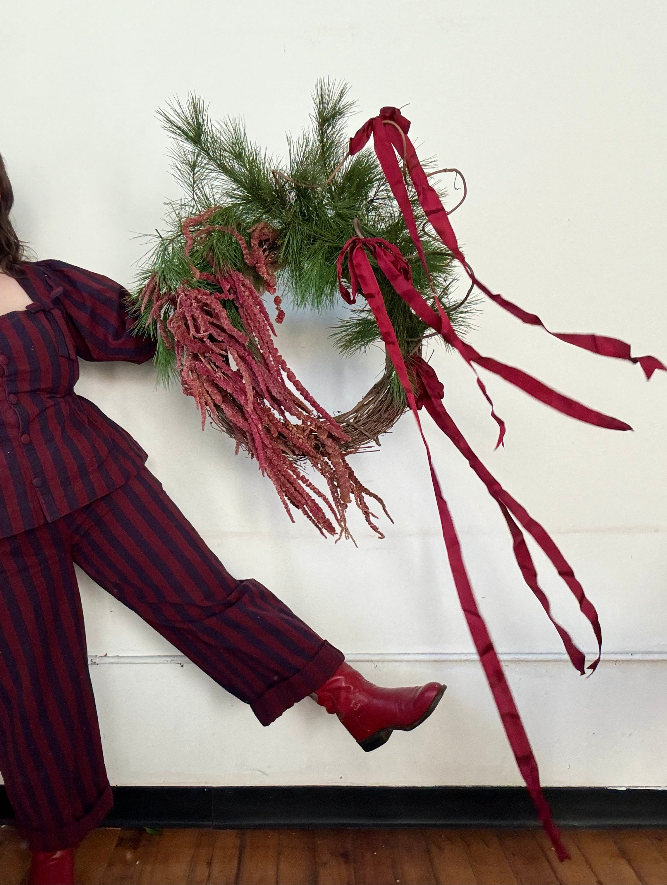 Maine florist in a red and blue striped suit and red boots holding a holiday wreath of maine grown amaranth and evergreens and ties in long silk ribbons