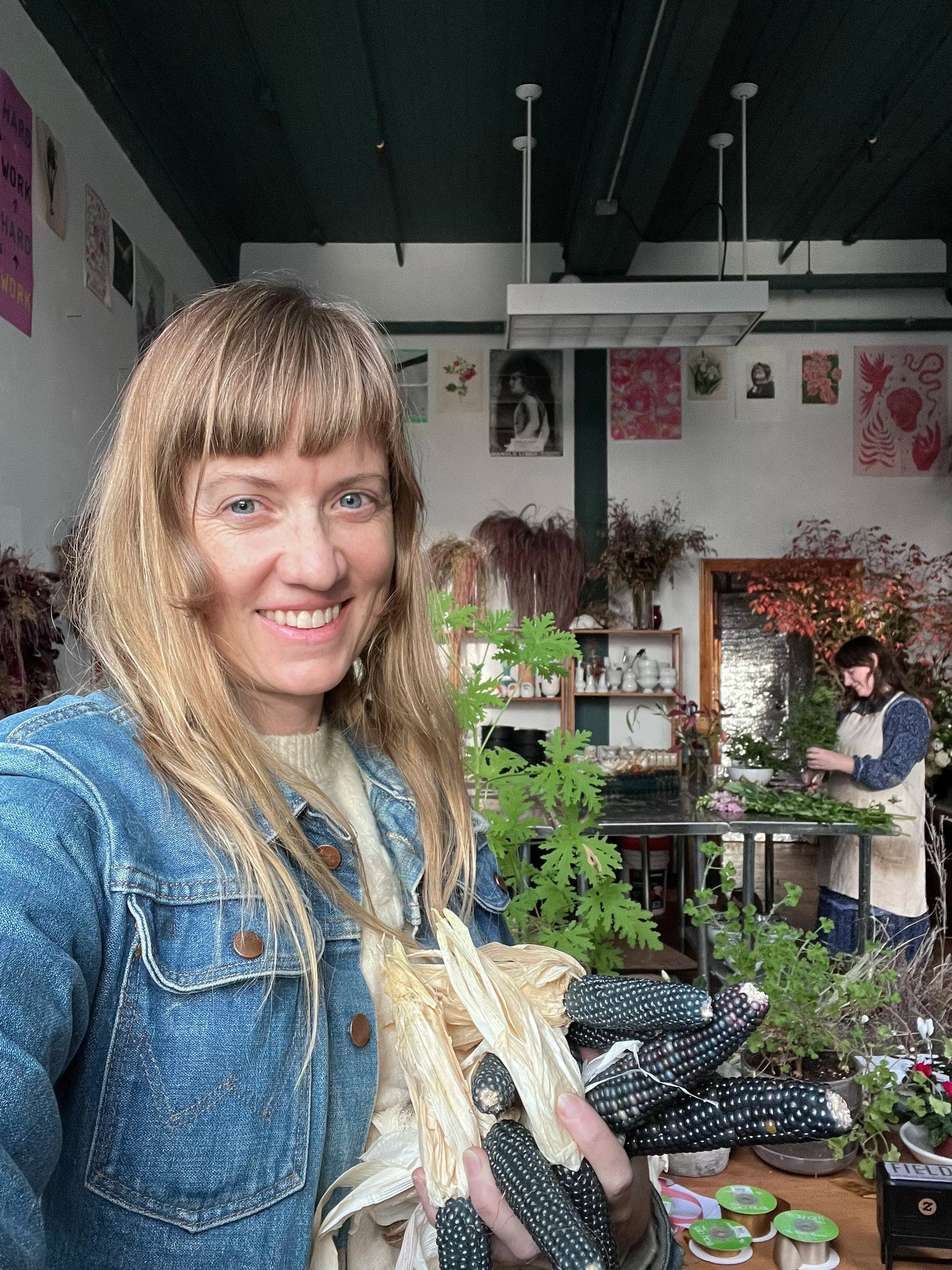 Woman holding a corn cob in an indoor setting with plants and people in the background