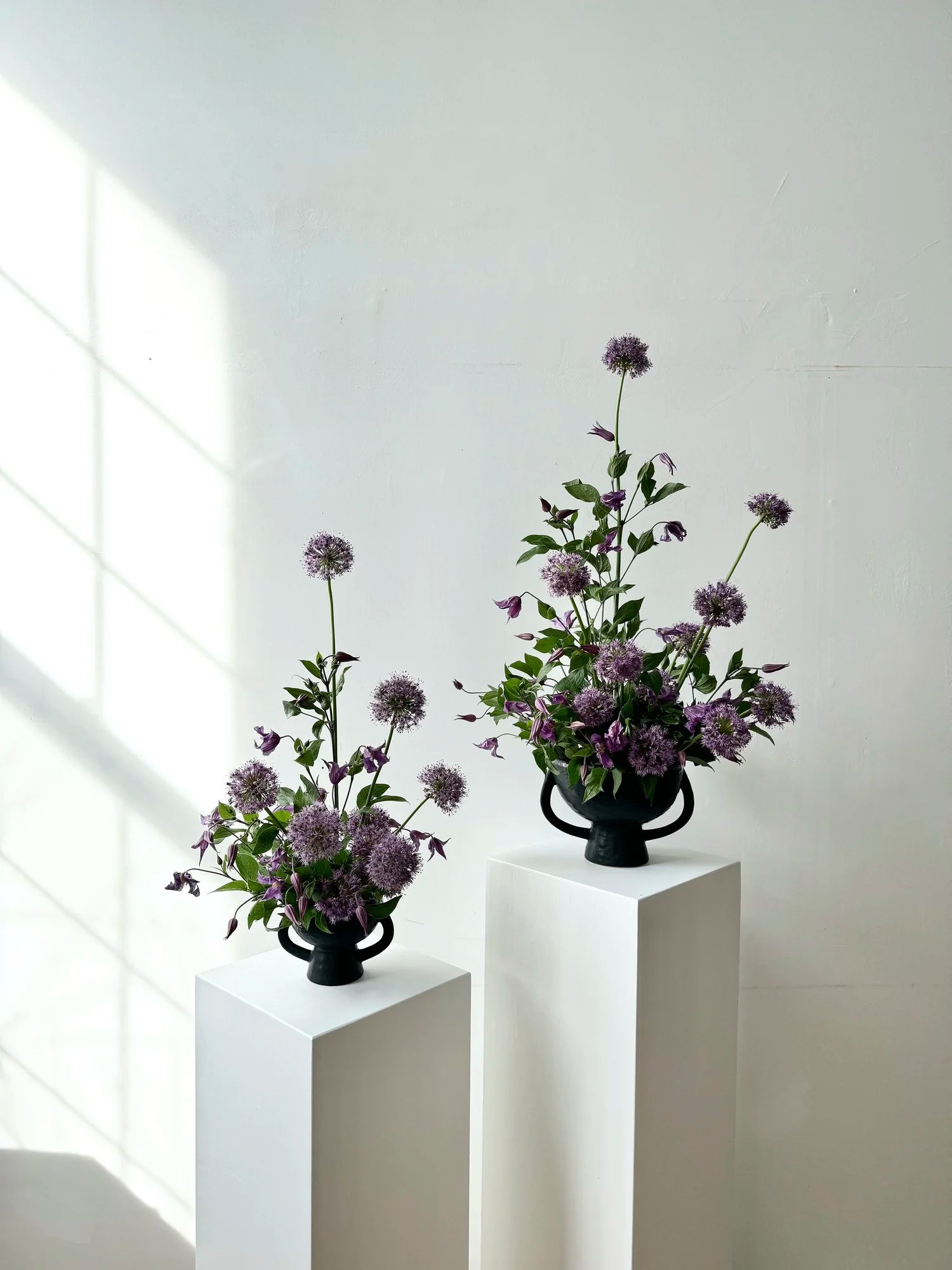 Two flower arrangements in black vases on white pedestals against a light gray wall.