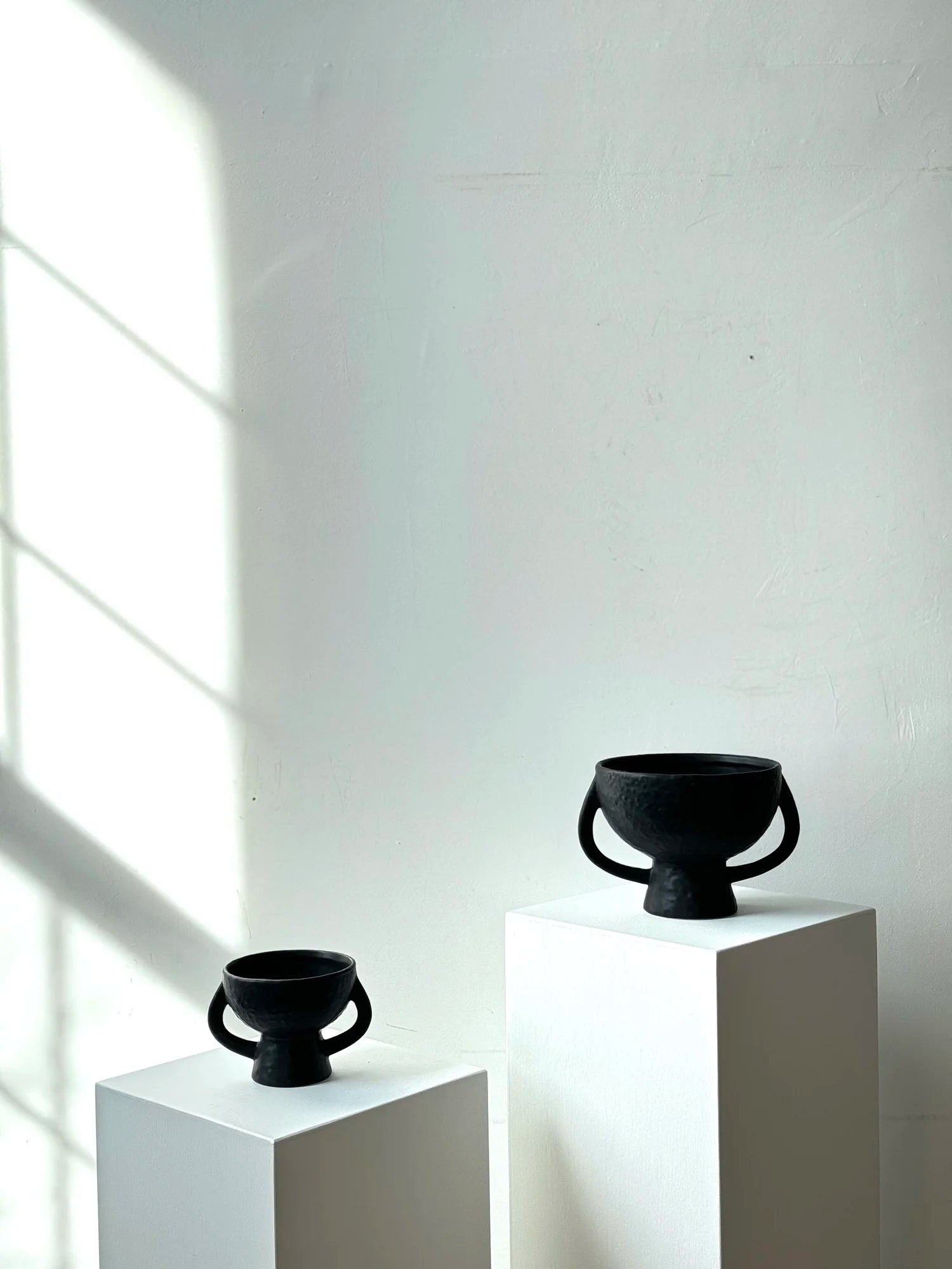 Two black ceramic vessels on white pedestals against a light gray wall.
