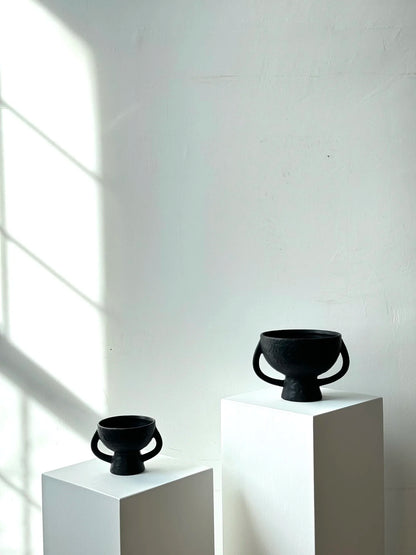 Two black ceramic vessels on white pedestals against a light gray wall.