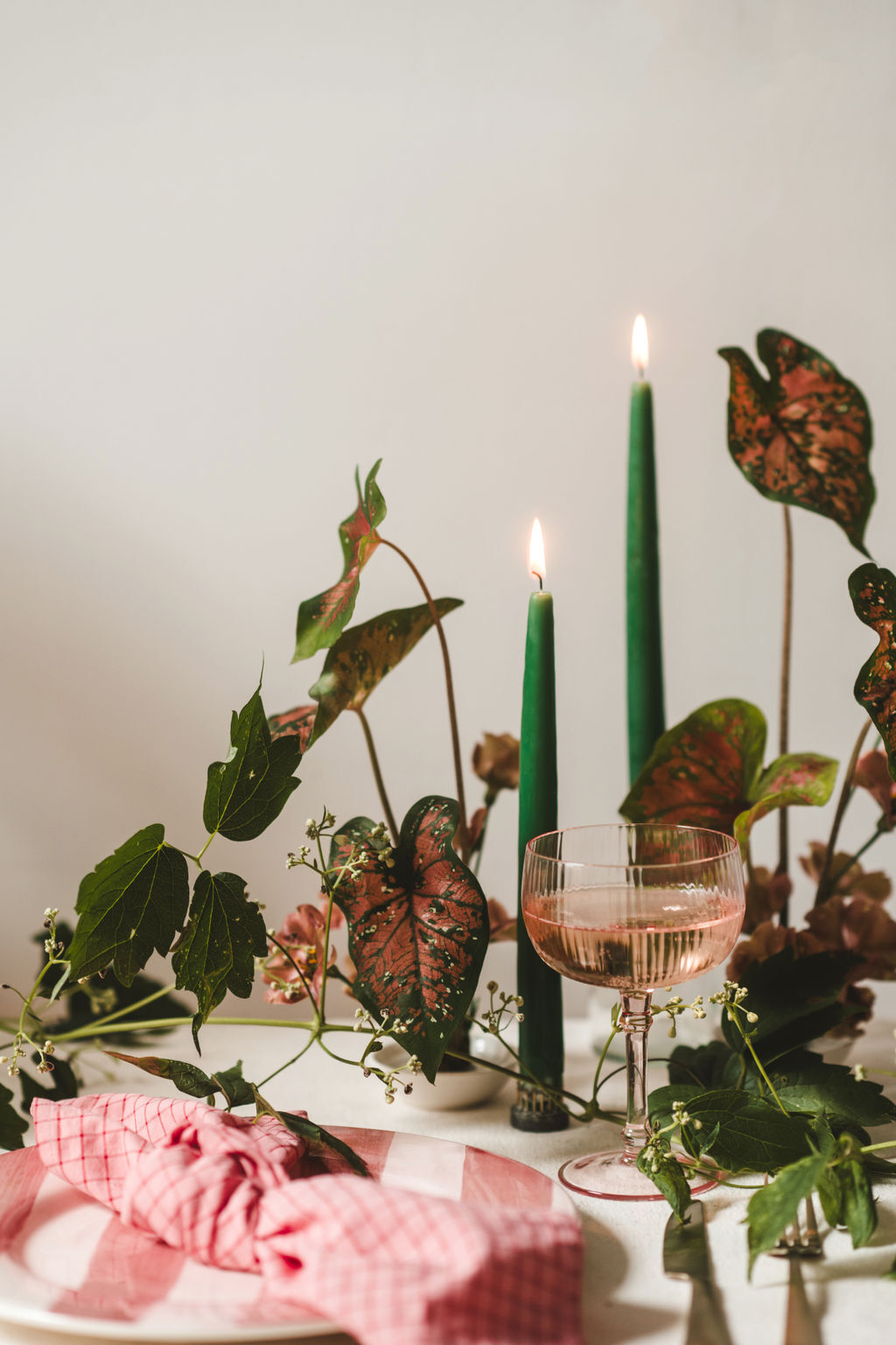 Decorative table setting with green candles, a pink glass, and leafy branches on a white surface.