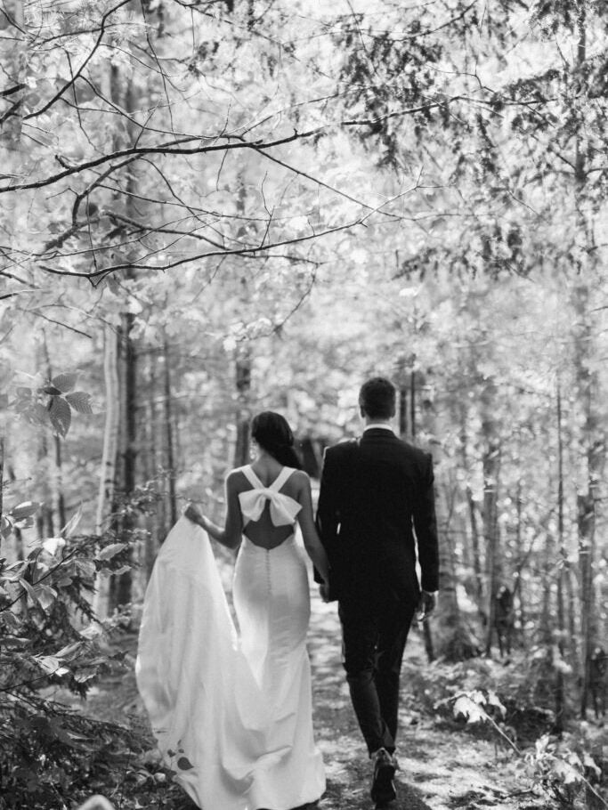Two people walking through a forest, one in a wedding dress with a bow back.