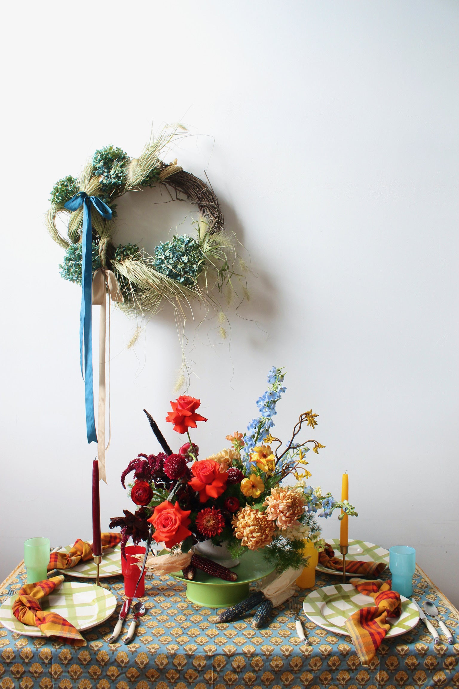 Decorative table setting with flowers, candles, and a wreath on a white wall background.