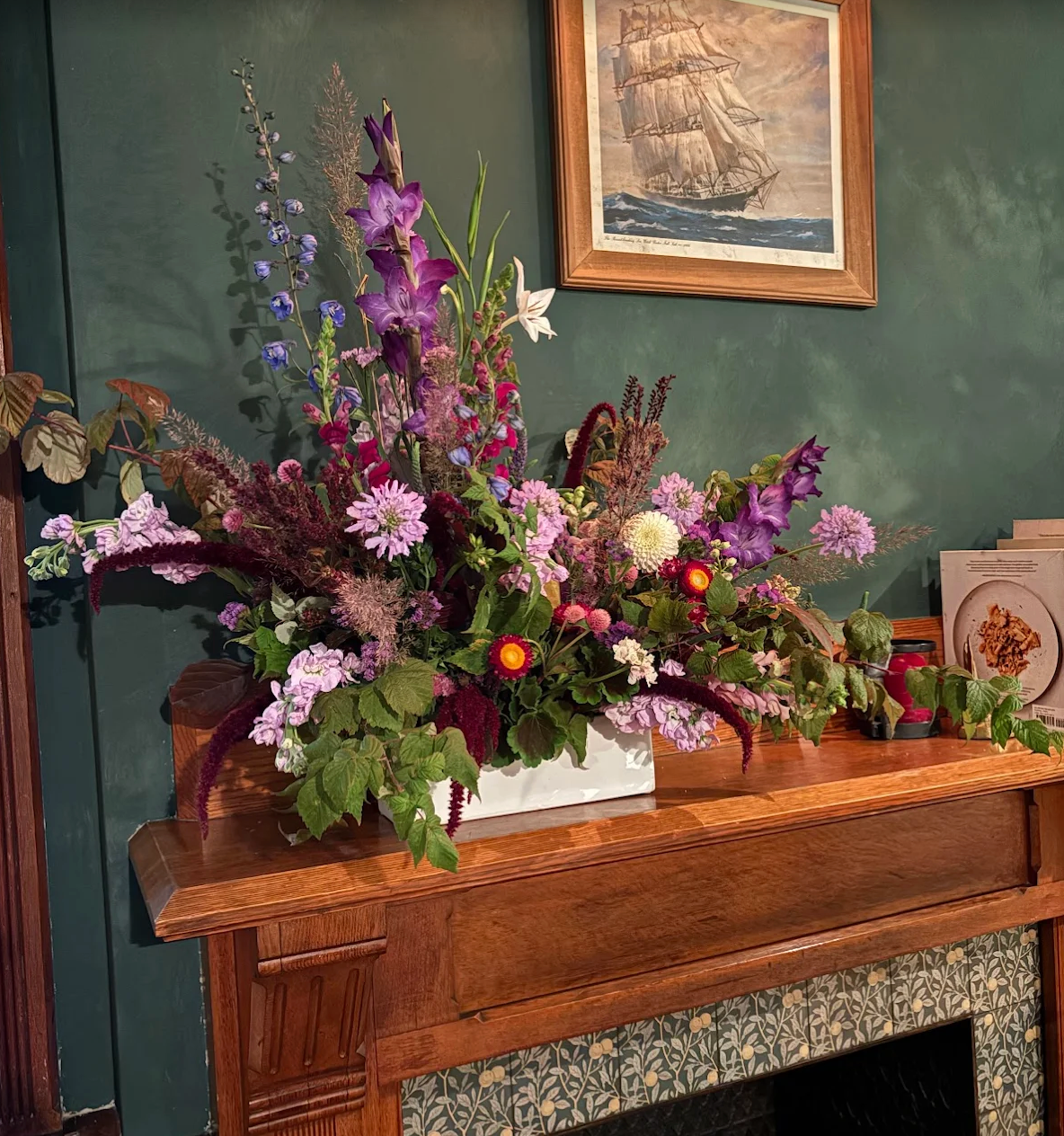 Floral arrangement on a wooden mantel with a framed picture of a ship on a green wall.