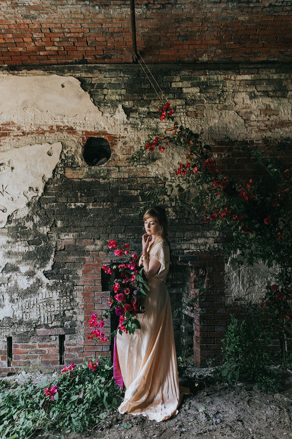 Woman in a flowing dress standing against a rustic brick wall with flowers.